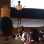 Front view of beautiful Asian businesswoman standing and giving presentation in front of the audience in auditorium