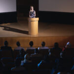 High view of blonde Caucasian businesswoman standing around podium and giving presentation to the audience in auditorium